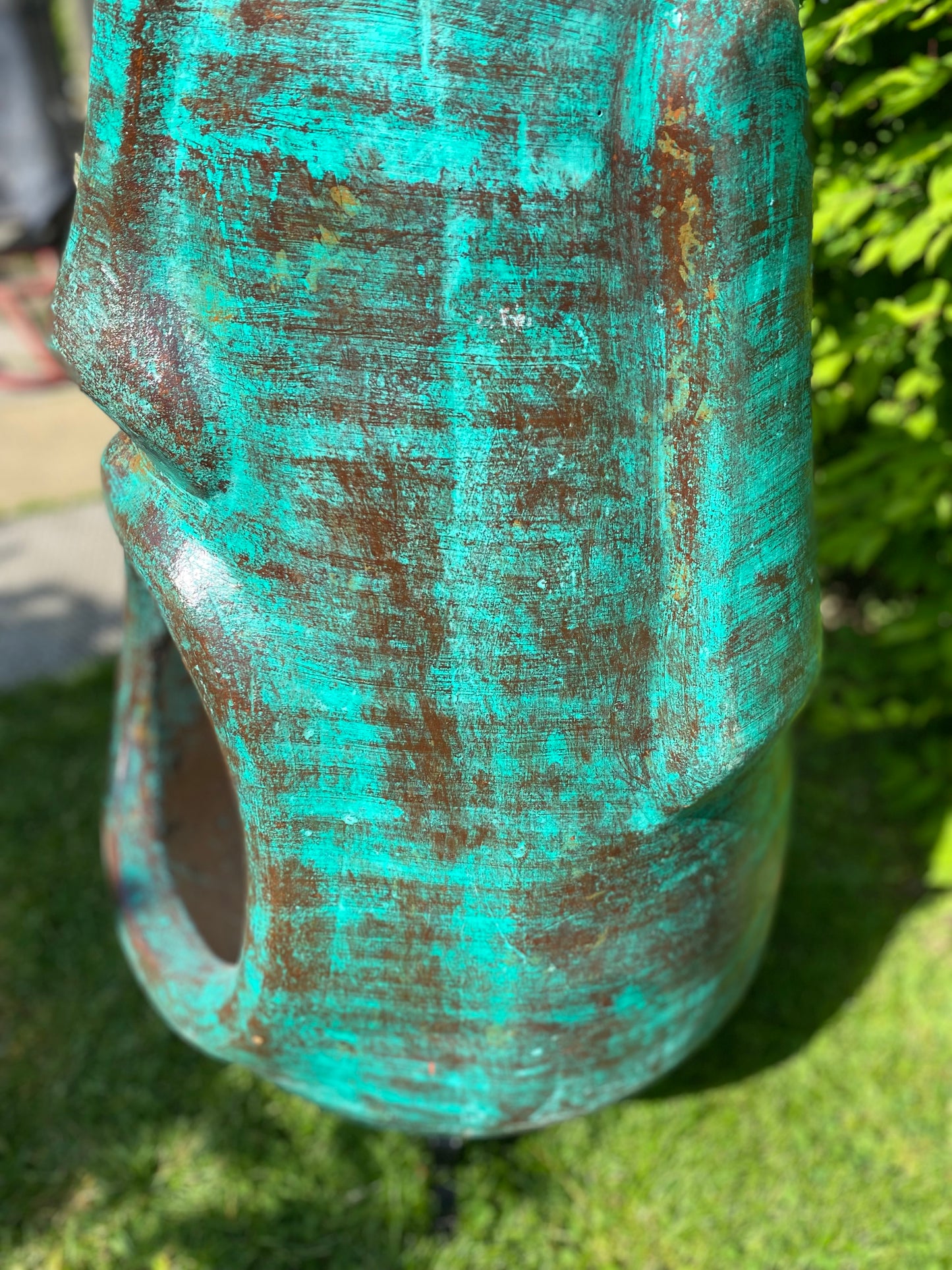 A rustic, rust-colored clay chiminea shaped like an Easter Island Moai head statue. The monolithic stone-face design features a prominent brow, a long straight nose, and a square chin. Flames flicker brightly from the wide, open mouth cavity, which serves as the fireplace opening. The outdoor heater sits on a black metal three-legged stand. close up