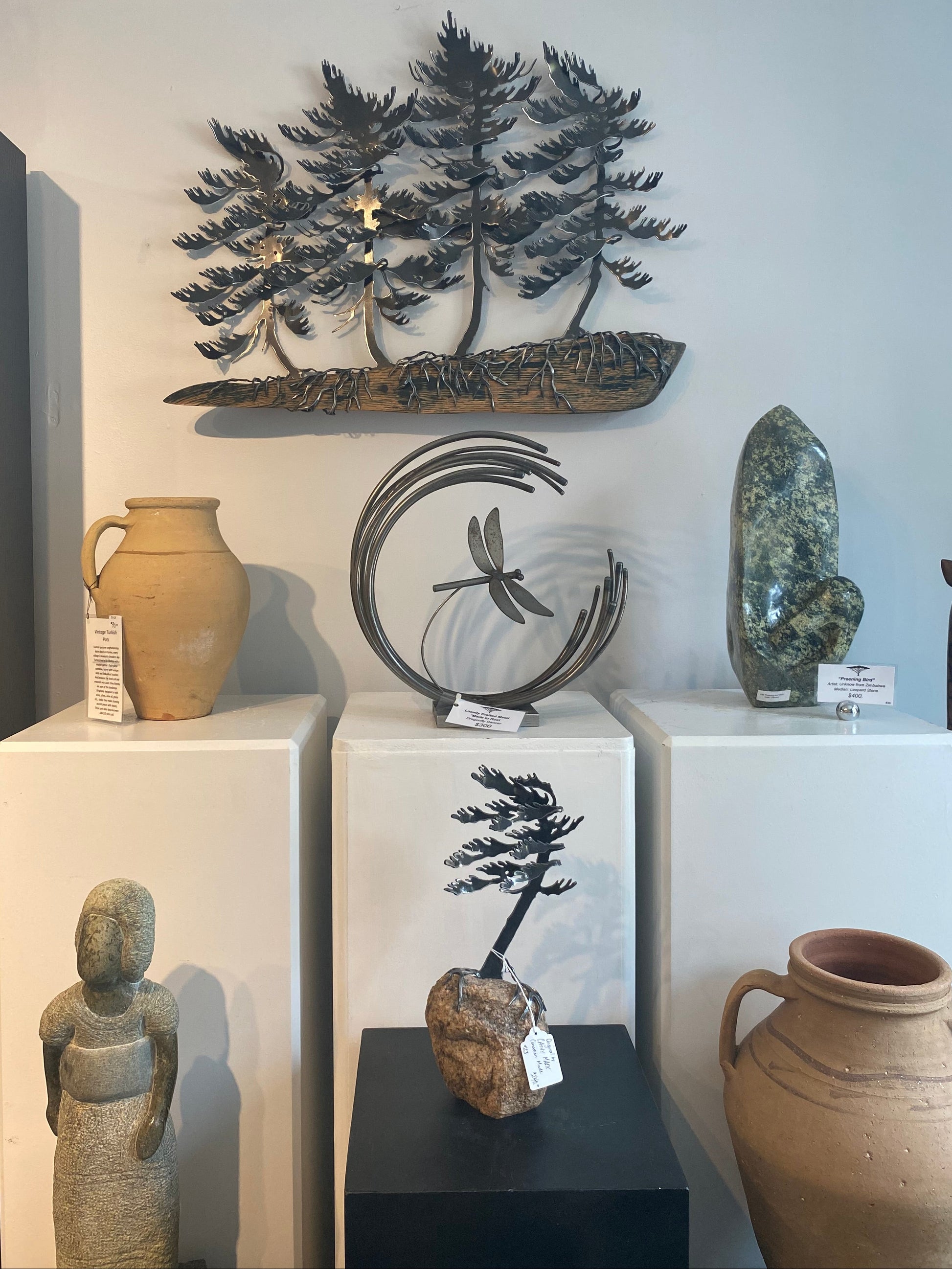 A collection of decorative wall art and sculptures displayed on white pedestals in a gallery setting, including metal tree sculptures, a dragonfly design, stone carvings, and ceramic vases.