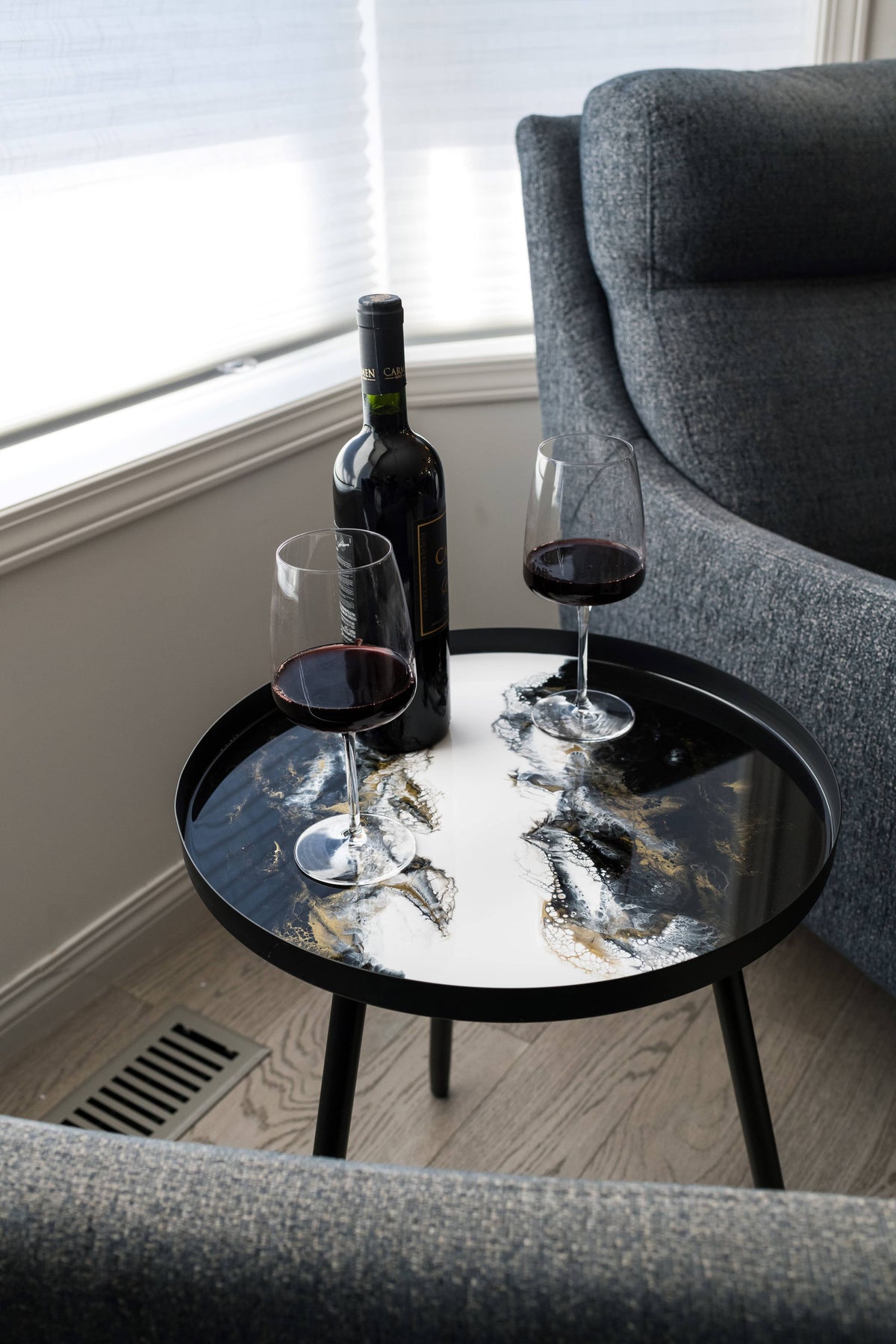 Black, white and gold resin side table with a wine bottle and two red wine glasses beside a grey armchair.