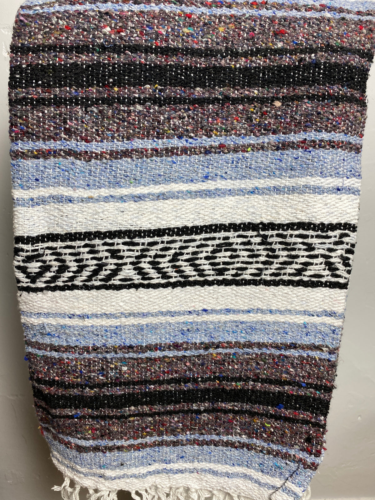 A woven textile blanket or throw with horizontal stripes in black, white, light blue, brown, and multicolored flecks, finished with a fringed edge. Falsa blanket from Mexico