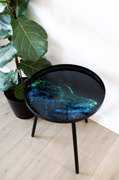 Black resin side table with flowing blue and green patterns, standing beside a leafy potted plant.
