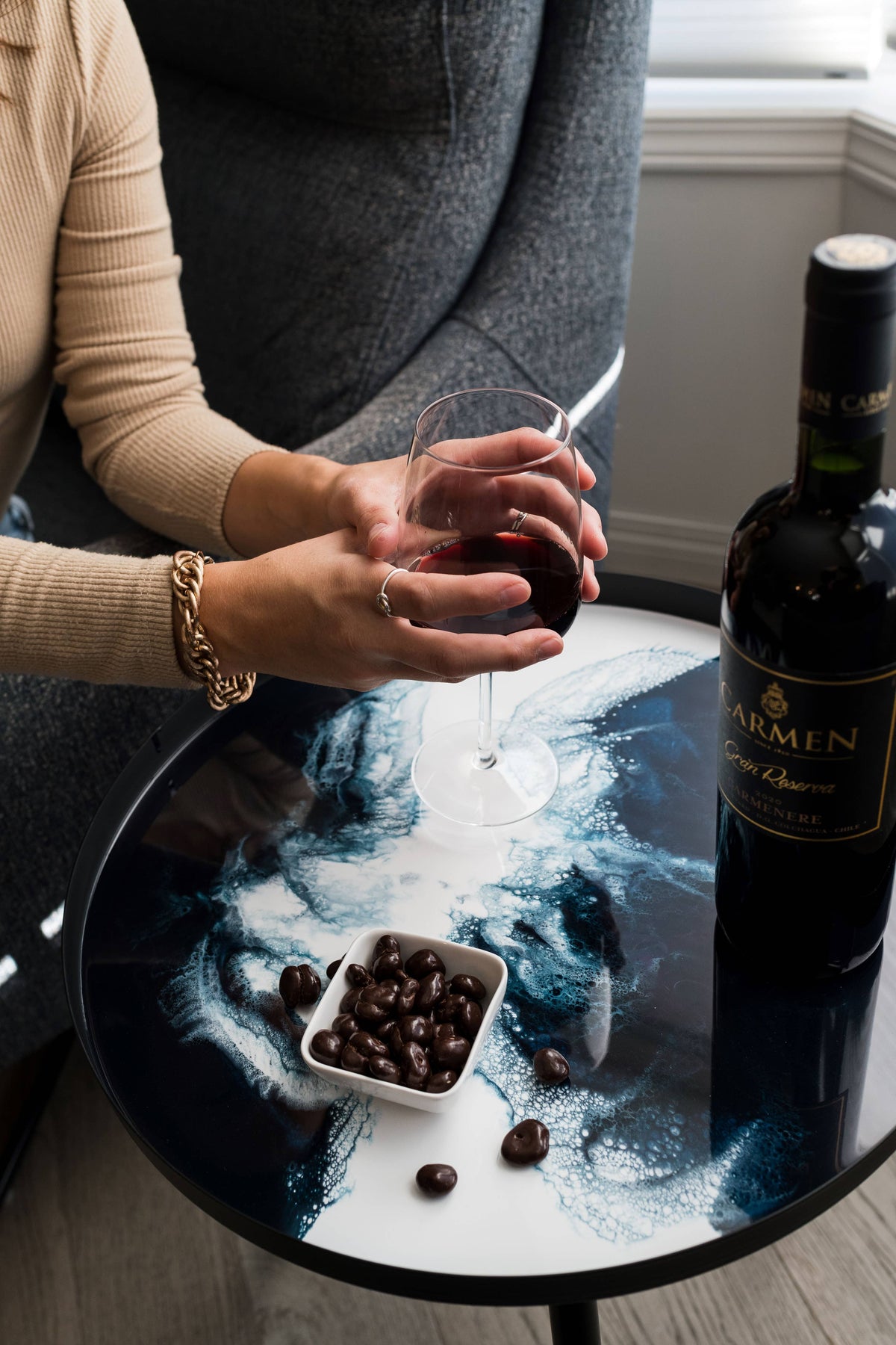 Close-up of a blue and white resin side table with a wine bottle and bowl of chocolates beside a person holding a wine glass.