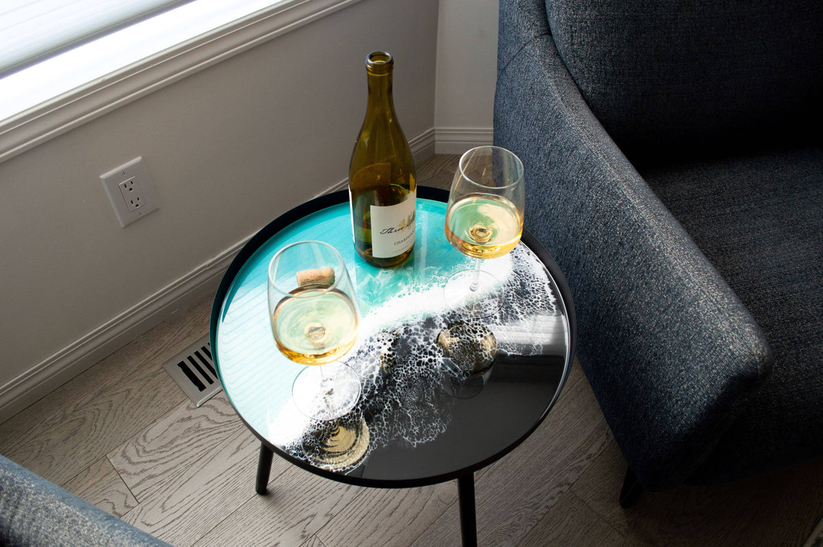 Overhead angled view of a turquoise and white resin side table holding a wine bottle and two glasses beside a grey chair.