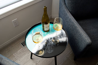 Overhead angled view of a turquoise and white resin side table holding a wine bottle and two glasses beside a grey chair.