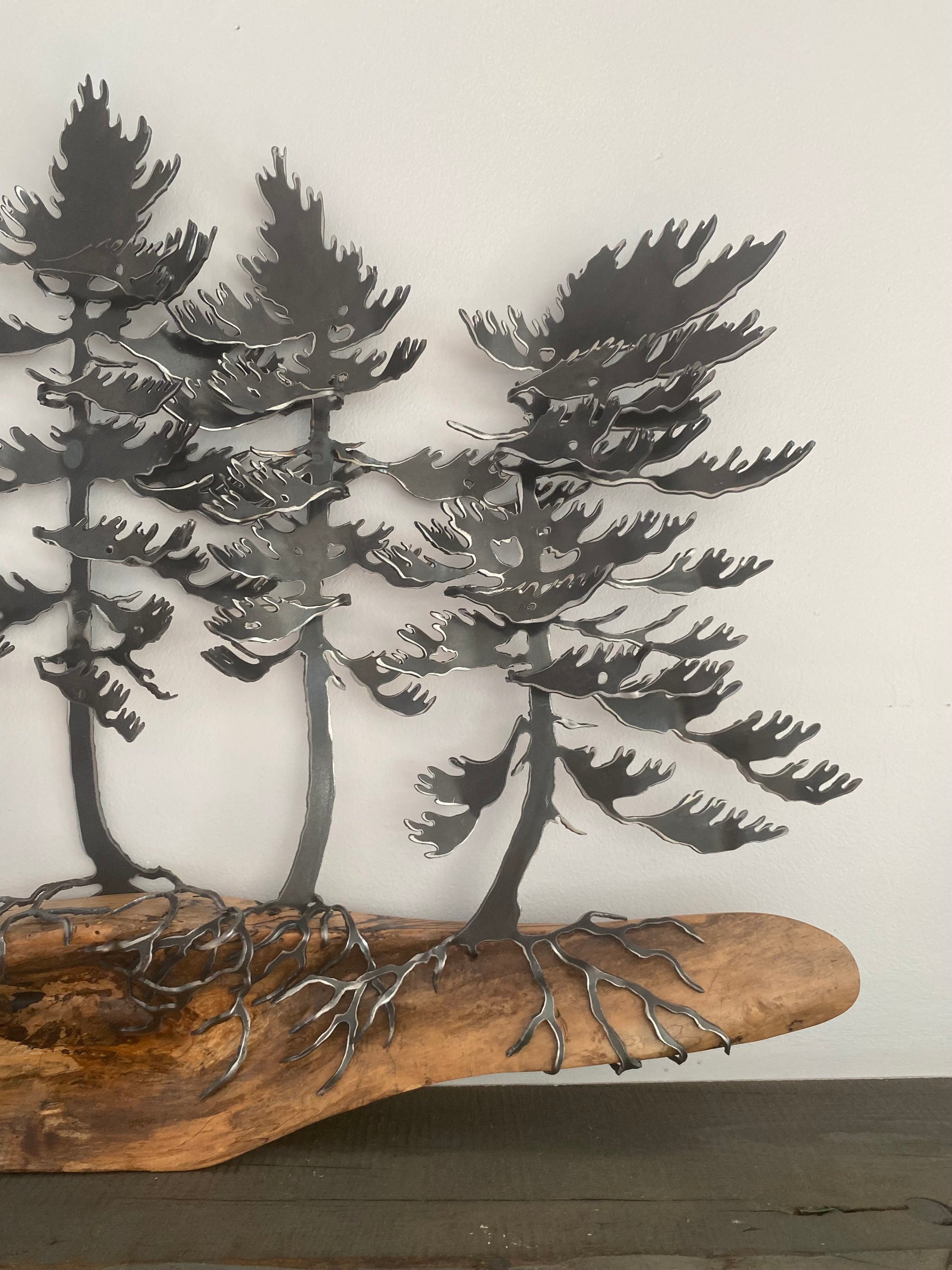 Metal wall art featuring three stylized pine trees with roots growing from a natural wood driftwood base, displayed against a plain off-white wall.