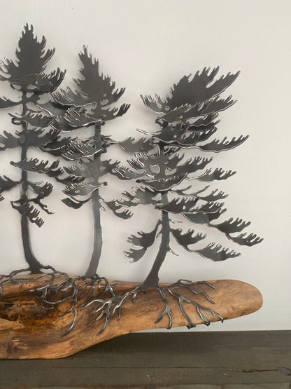 Metal wall art featuring three stylized pine trees with roots growing from a natural wood driftwood base, displayed against a plain off-white wall.