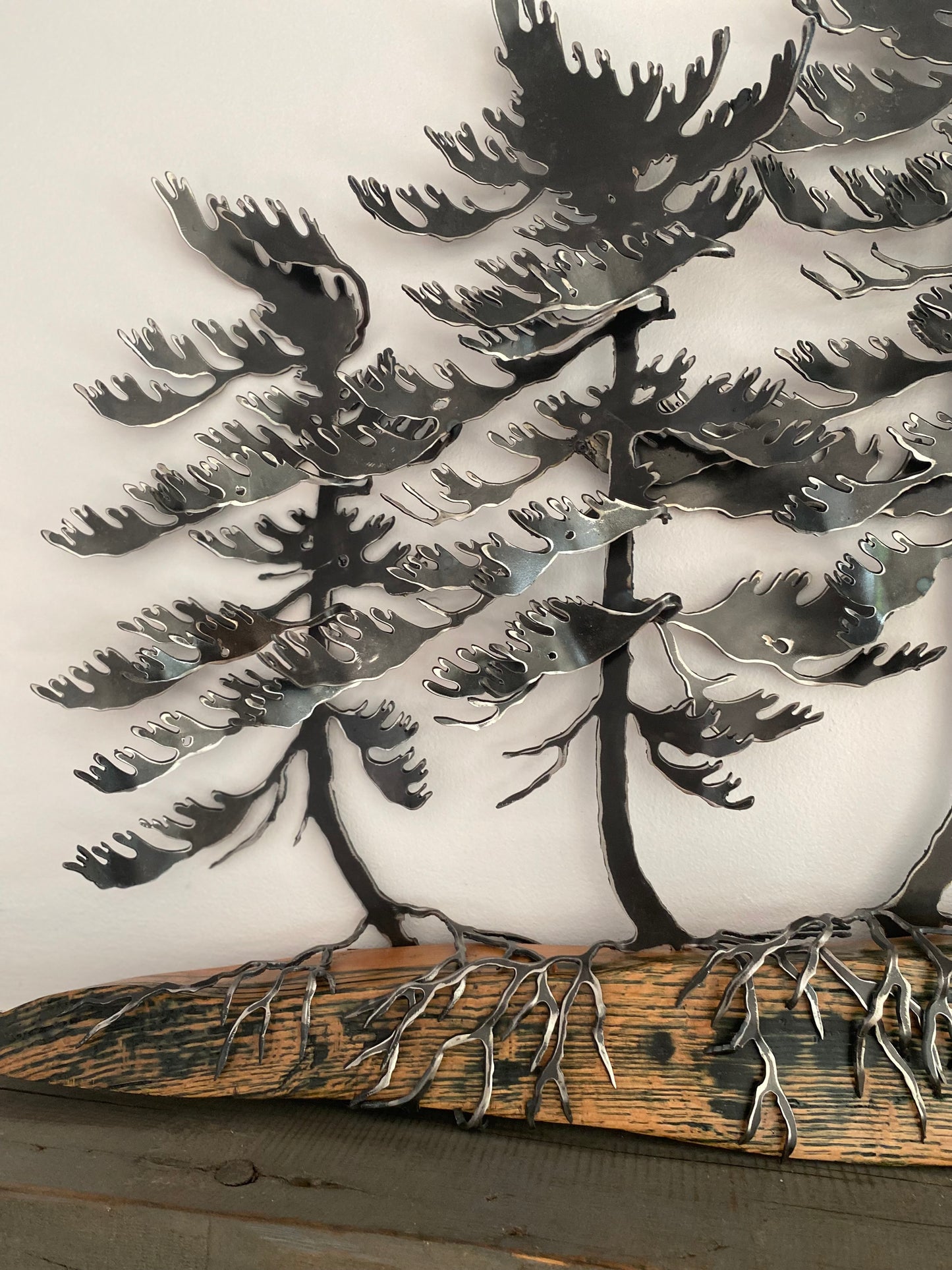 Metallic tree wall art featuring layered pine tree silhouettes with exposed roots on a rustic wood base, photographed against a plain light background.