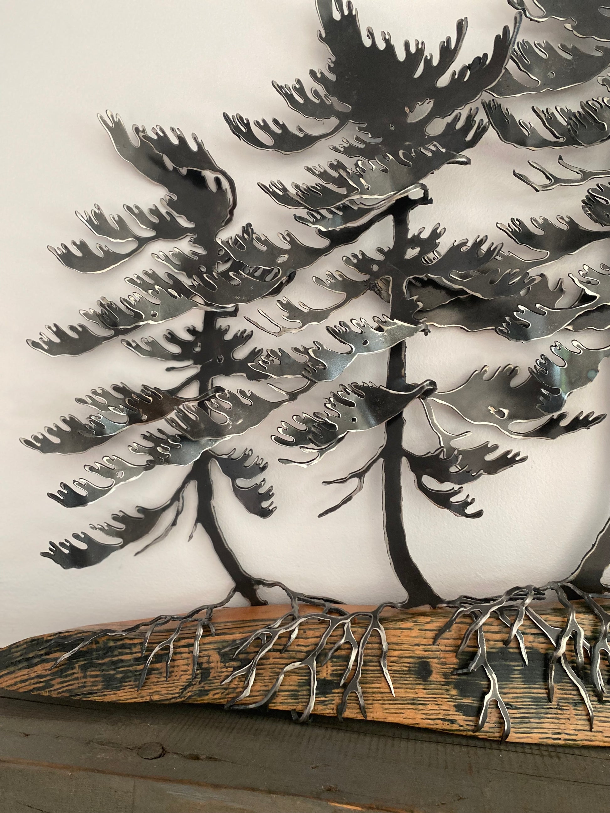Metallic tree wall art featuring layered pine tree silhouettes with exposed roots on a rustic wood base, photographed against a plain light background.