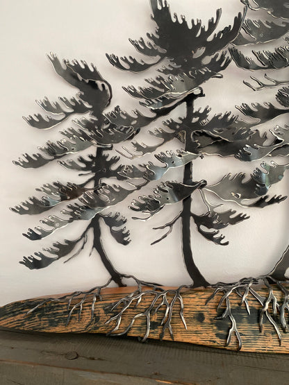 Metallic tree wall art featuring layered pine tree silhouettes with exposed roots on a rustic wood base, photographed against a plain light background.