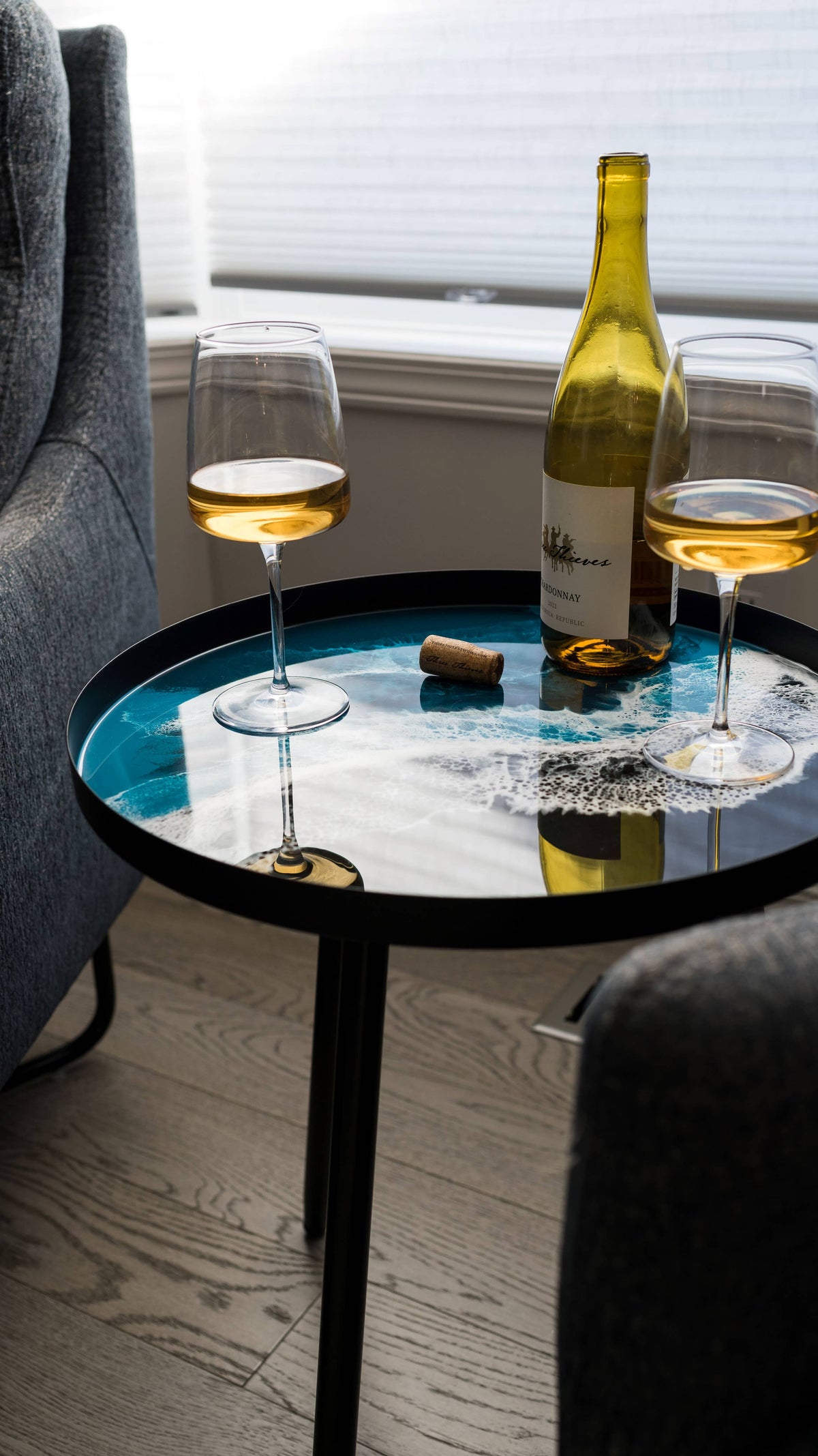 Turquoise ocean-pattern resin side table holding a wine bottle and two glasses between grey upholstered chairs.