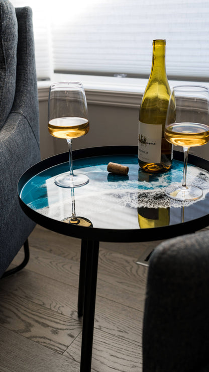 Turquoise ocean-pattern resin side table holding a wine bottle and two glasses between grey upholstered chairs.
