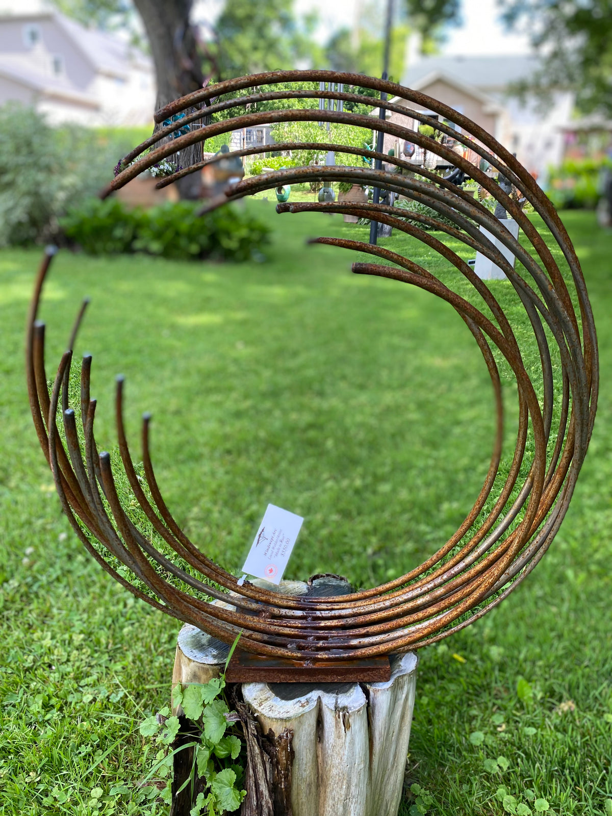 Abstract metal sculpture made of rusted curved tubes, displayed on a tree stump in a grassy backyard.