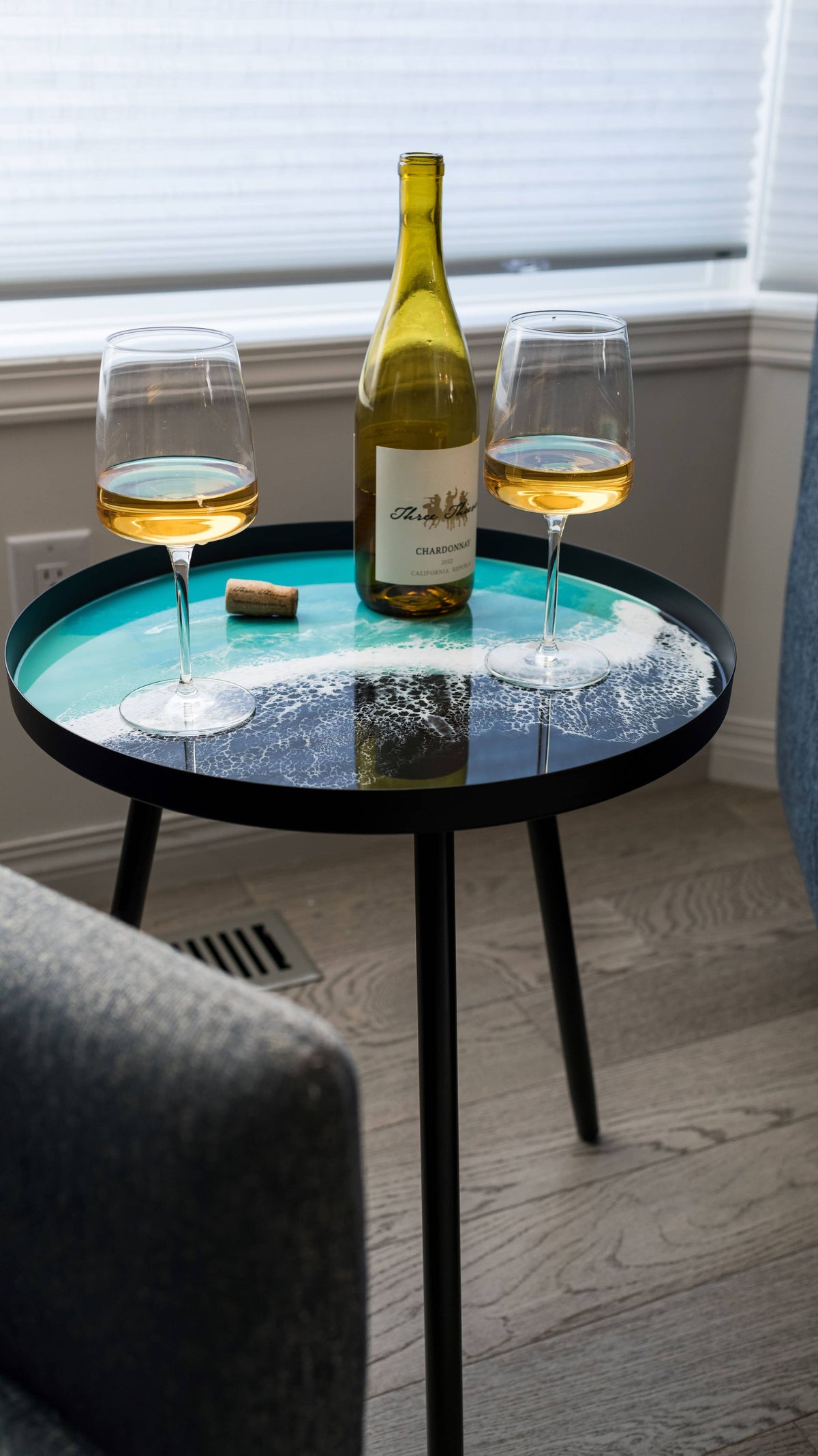 Turquoise and white resin side table with black metal legs, styled with a wine bottle and two glasses beside an armchair.