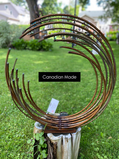 Rustic metal sculpture made of multiple curved, weathered brown metal rods arranged in a spiral shape, displayed on a tree stump in a grassy backyard with a 'Canadian Made' label overlay.