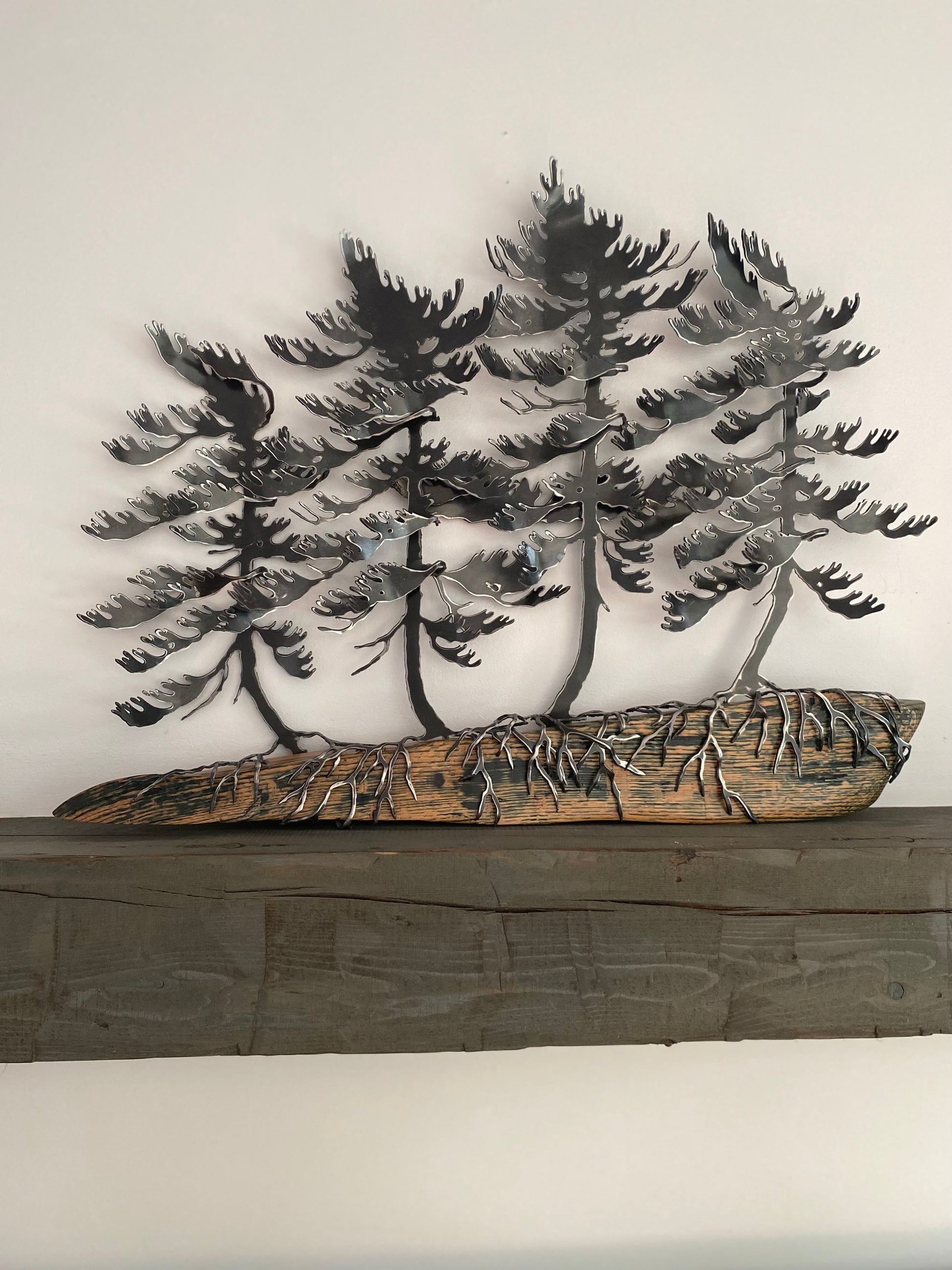 Metallic black pine tree wall art mounted above a rustic dark wood mantel against a plain off-white wall.