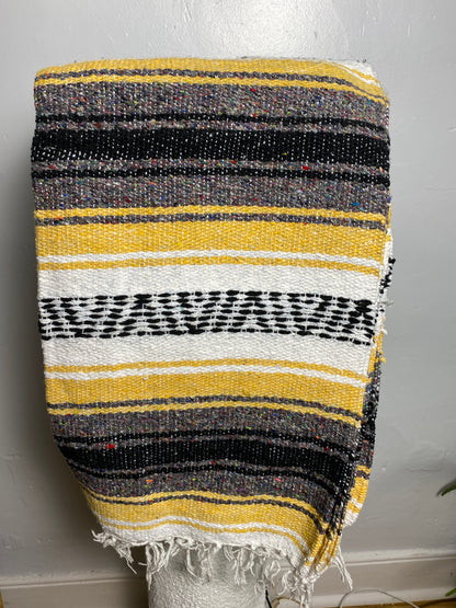 A woven striped blanket or throw with a fringed edge, displayed on a white stand against a light gray wall. The pattern features horizontal stripes of yellow, black, white, and gray. Falsa blanket from Mexico
