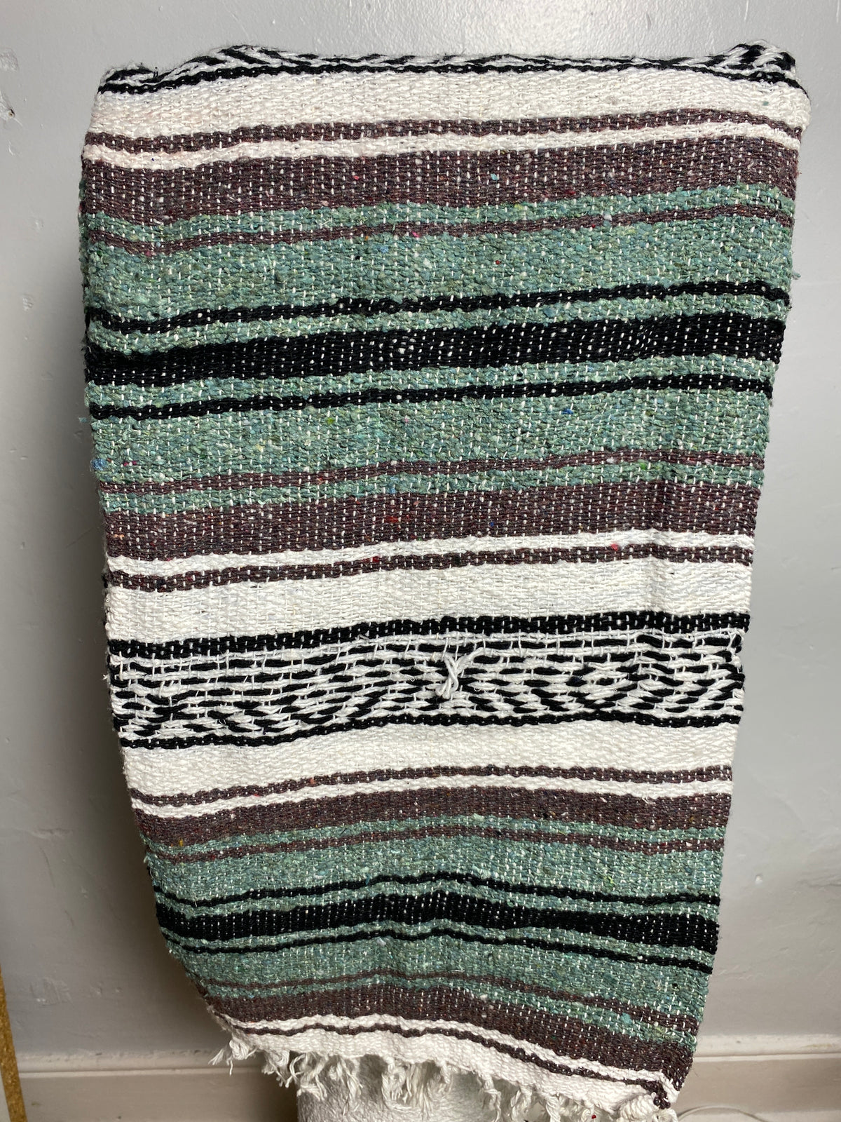 A woven textile blanket or throw with horizontal stripes in sage green, black, white, and taupe, featuring a fringed edge and a geometric patterned band, Falsa blanket from Mexico