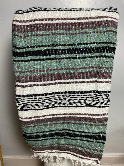 A woven textile blanket or throw with horizontal stripes in sage green, black, white, and taupe, featuring a fringed edge and a geometric patterned band, Falsa blanket from Mexico