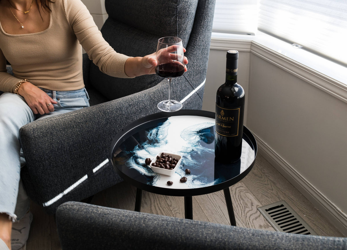 Blue and white resin side table styled with wine and chocolates beside a person seated in a grey armchair.