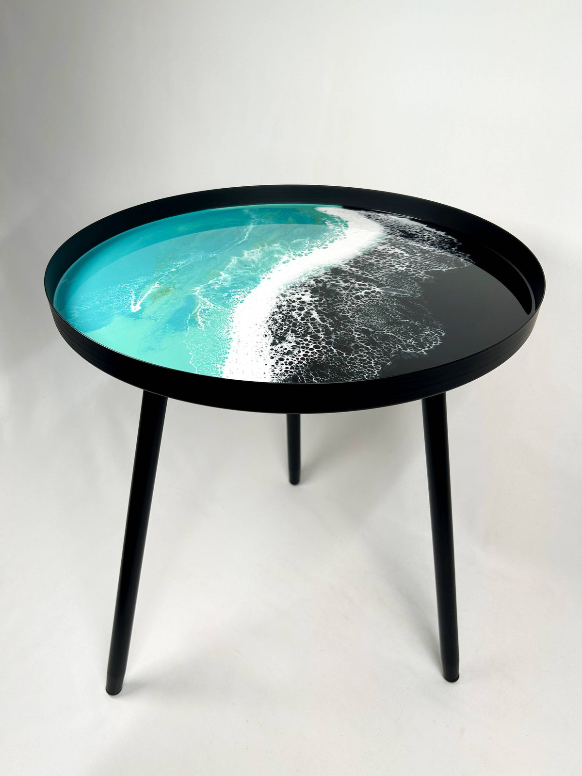 Turquoise and white wave-pattern resin tabletop with a black rim and three black metal legs on a plain background.