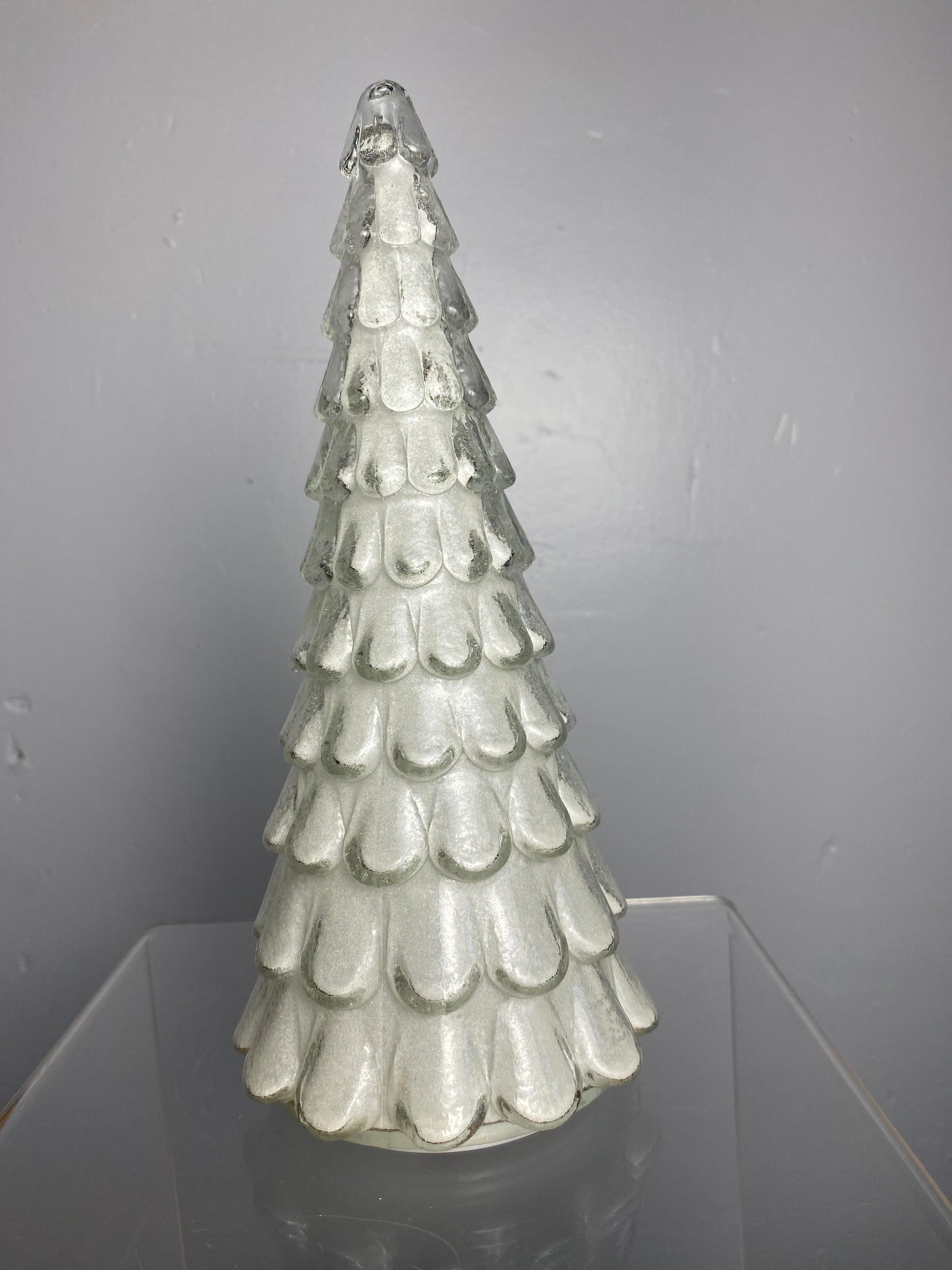 A clear glass Christmas tree ornament shaped like a tiered cone, decorated with white scalloped glass scales, shown on a neutral gray background.