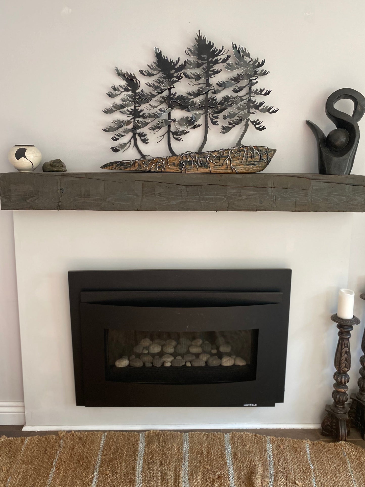 A rustic dark gray stone fireplace mantel decorated with a metal tree sculpture, a carved log, and a black bird figurine, set above a black electric fireplace insert.