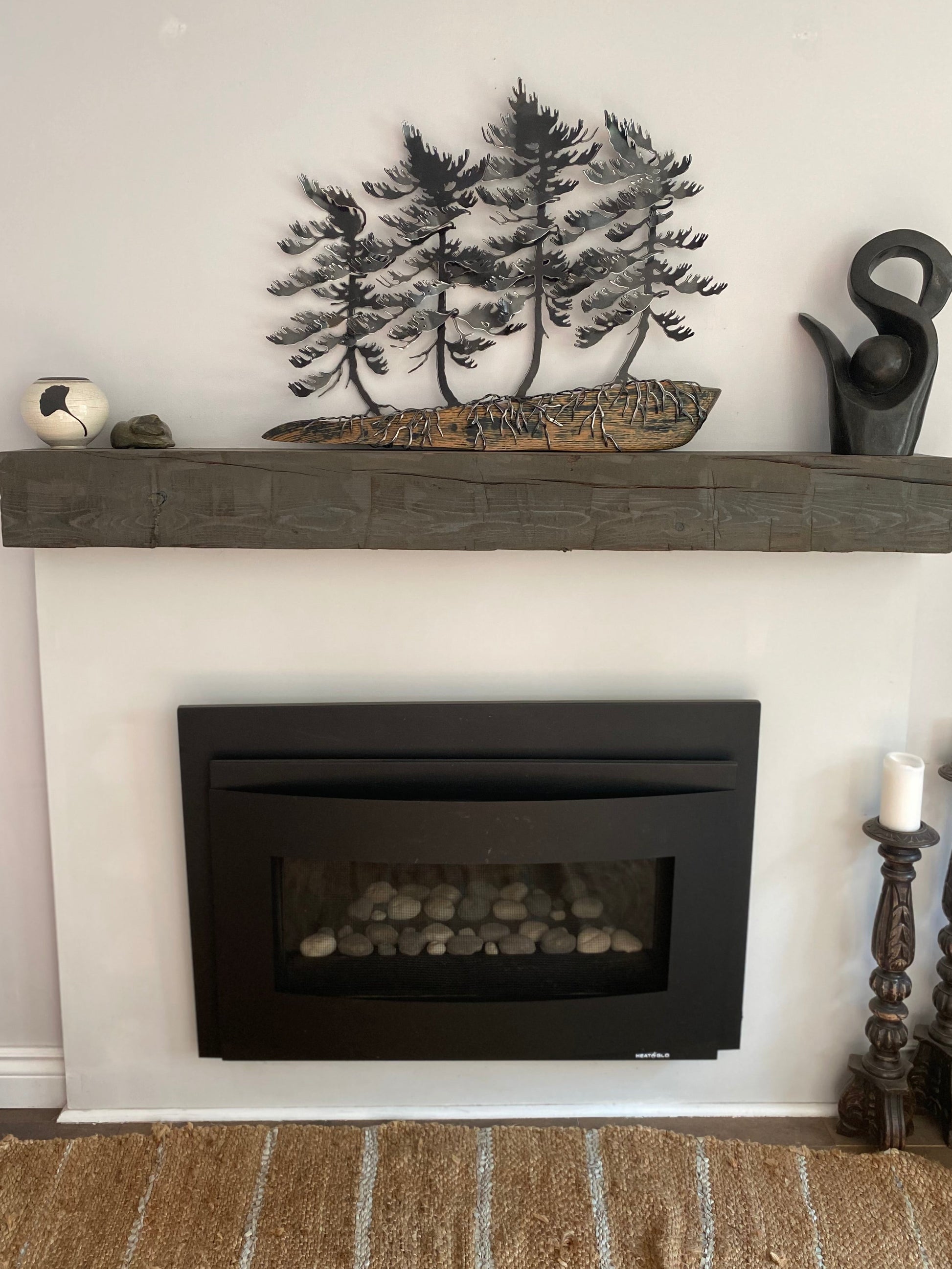 A rustic dark gray stone fireplace mantel decorated with a metal tree sculpture, a carved log, and a black bird figurine, set above a black electric fireplace insert.
