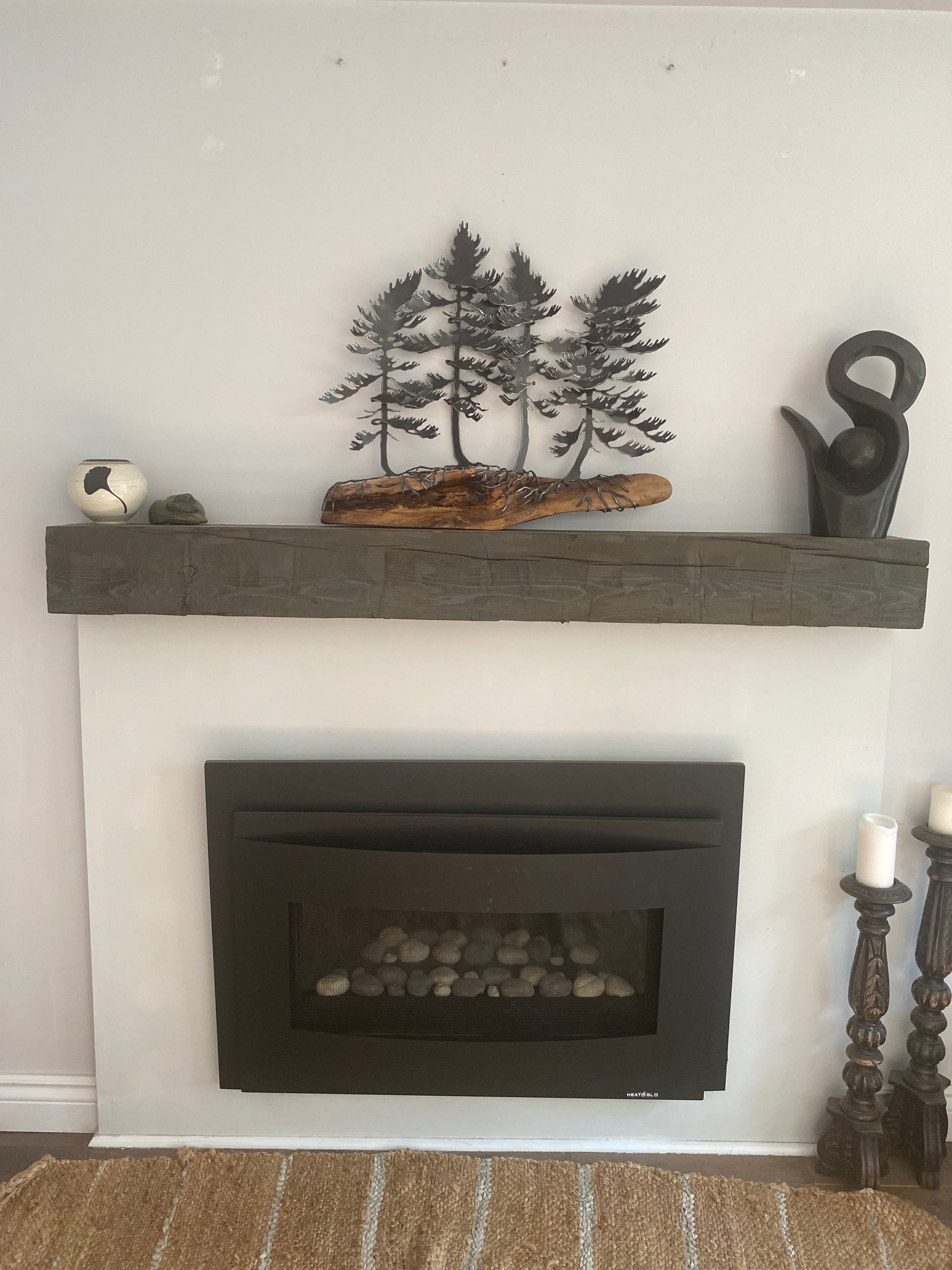 A dark wood mantel shelf with a textured finish, styled with a black metal tree sculpture, a piece of driftwood, and a small silver bell, shown next to a black fireplace insert on a white wall.