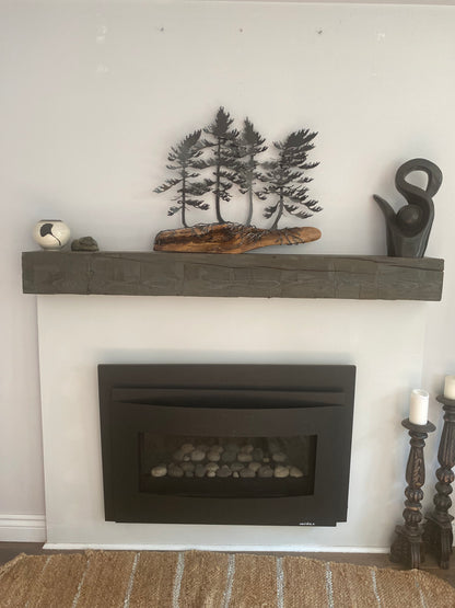 A dark wood mantel shelf with a textured finish, styled with a black metal tree sculpture, a piece of driftwood, and a small silver bell, shown next to a black fireplace insert on a white wall.