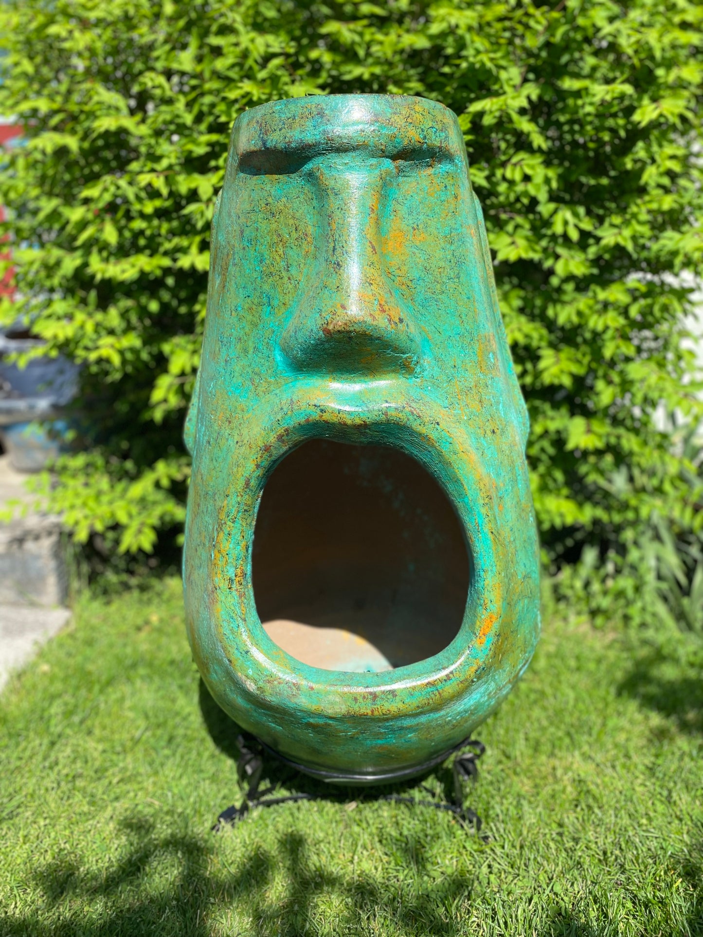 A rustic, rust-colored clay chiminea shaped like an Easter Island Moai head statue. The monolithic stone-face design features a prominent brow, a long straight nose, and a square chin. Flames flicker brightly from the wide, open mouth cavity, which serves as the fireplace opening. The outdoor heater sits on a black metal three-legged stand.