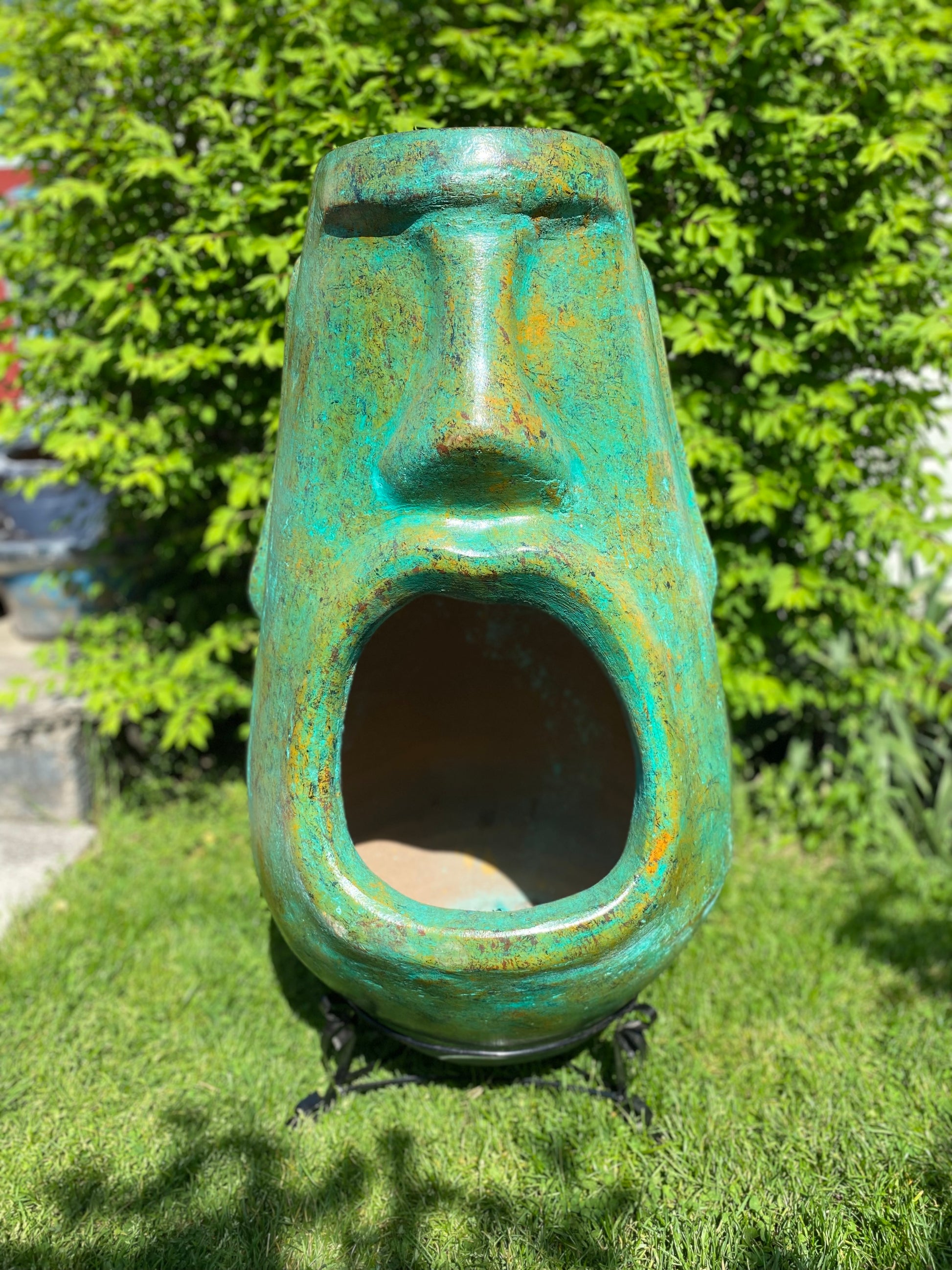 A rustic, rust-colored clay chiminea shaped like an Easter Island Moai head statue. The monolithic stone-face design features a prominent brow, a long straight nose, and a square chin. Flames flicker brightly from the wide, open mouth cavity, which serves as the fireplace opening. The outdoor heater sits on a black metal three-legged stand.