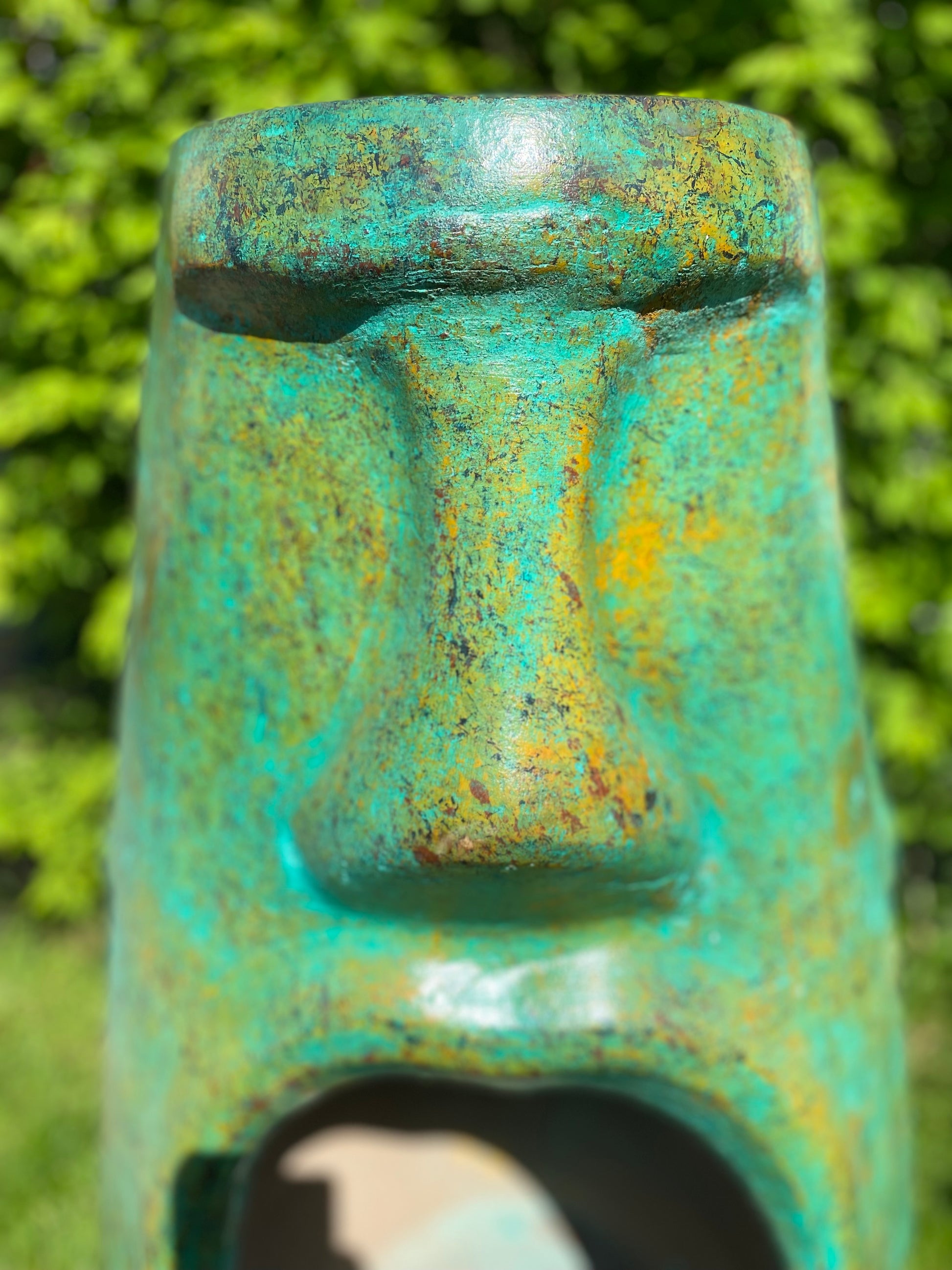 A rustic, rust-colored clay chiminea shaped like an Easter Island Moai head statue. The monolithic stone-face design features a prominent brow, a long straight nose, and a square chin. Flames flicker brightly from the wide, open mouth cavity, which serves as the fireplace opening. The outdoor heater sits on a black metal three-legged stand. close up