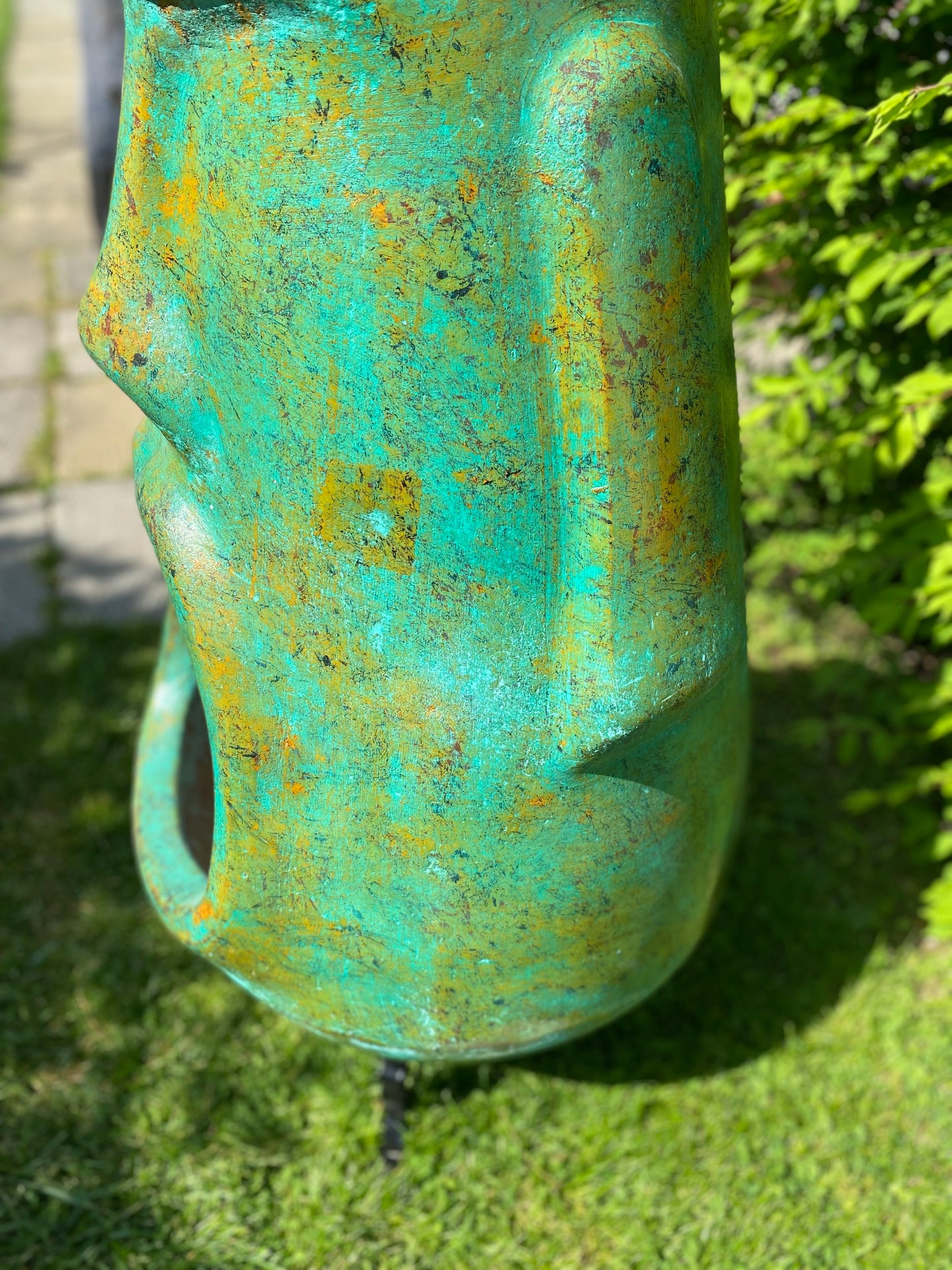 A rustic, rust-colored clay chiminea shaped like an Easter Island Moai head statue. The monolithic stone-face design features a prominent brow, a long straight nose, and a square chin. Flames flicker brightly from the wide, open mouth cavity, which serves as the fireplace opening. The outdoor heater sits on a black metal three-legged stand. Close up of green colour