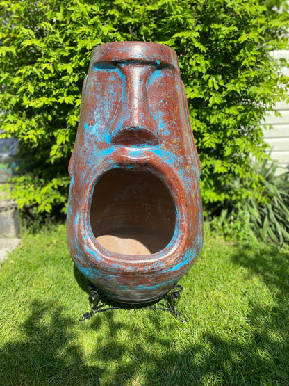 A rustic, rust-colored clay chiminea shaped like an Easter Island Moai head statue. The monolithic stone-face design features a prominent brow, a long straight nose, and a square chin. Flames flicker brightly from the wide, open mouth cavity, which serves as the fireplace opening. The outdoor heater sits on a black metal three-legged stand.