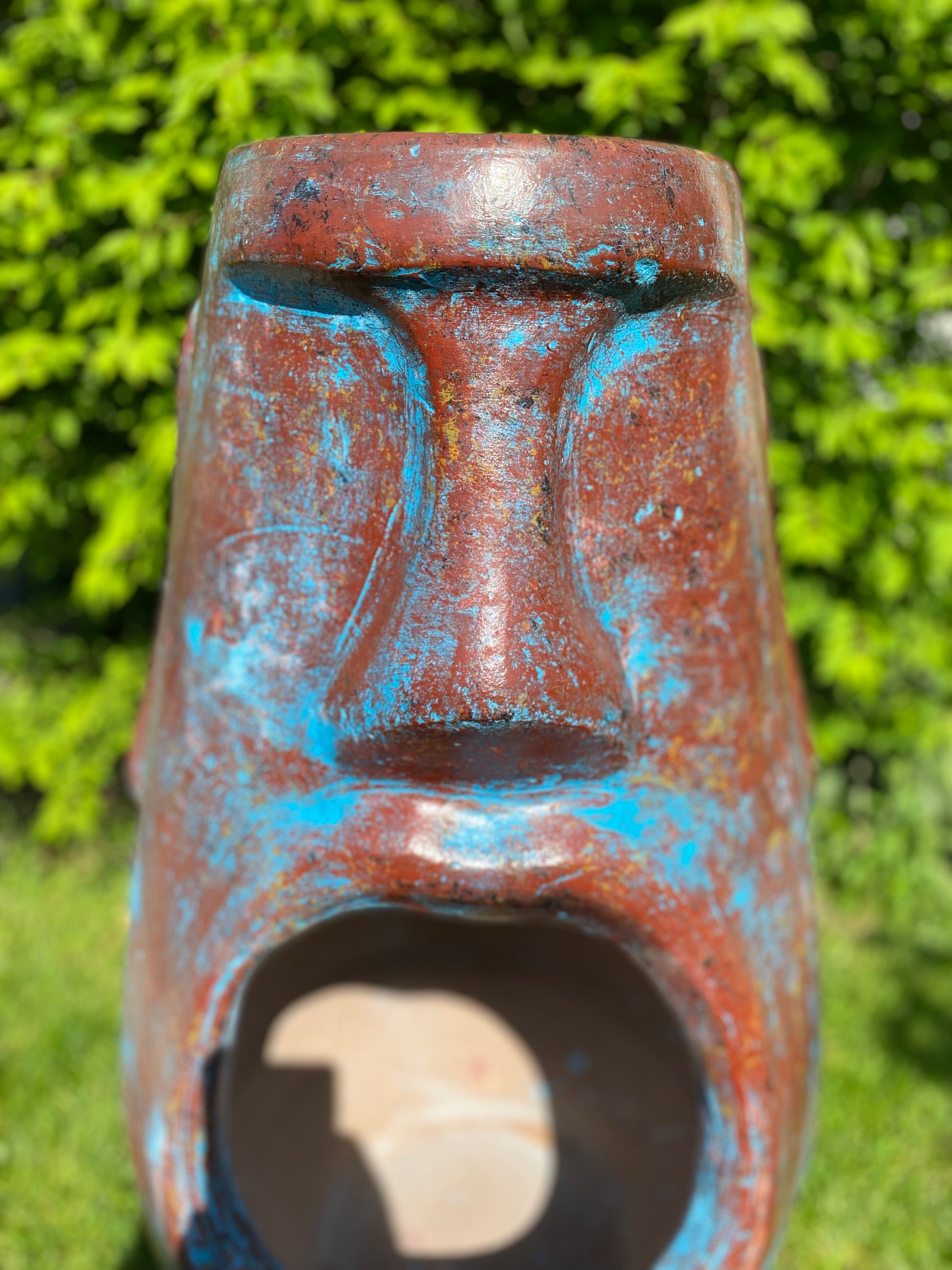 A rustic, rust-colored clay chiminea shaped like an Easter Island Moai head statue. The monolithic stone-face design features a prominent brow, a long straight nose, and a square chin. Flames flicker brightly from the wide, open mouth cavity, which serves as the fireplace opening. The outdoor heater sits on a black metal three-legged stand. close up