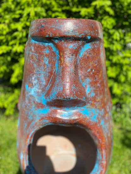 A rustic, rust-colored clay chiminea shaped like an Easter Island Moai head statue. The monolithic stone-face design features a prominent brow, a long straight nose, and a square chin. Flames flicker brightly from the wide, open mouth cavity, which serves as the fireplace opening. The outdoor heater sits on a black metal three-legged stand. close up