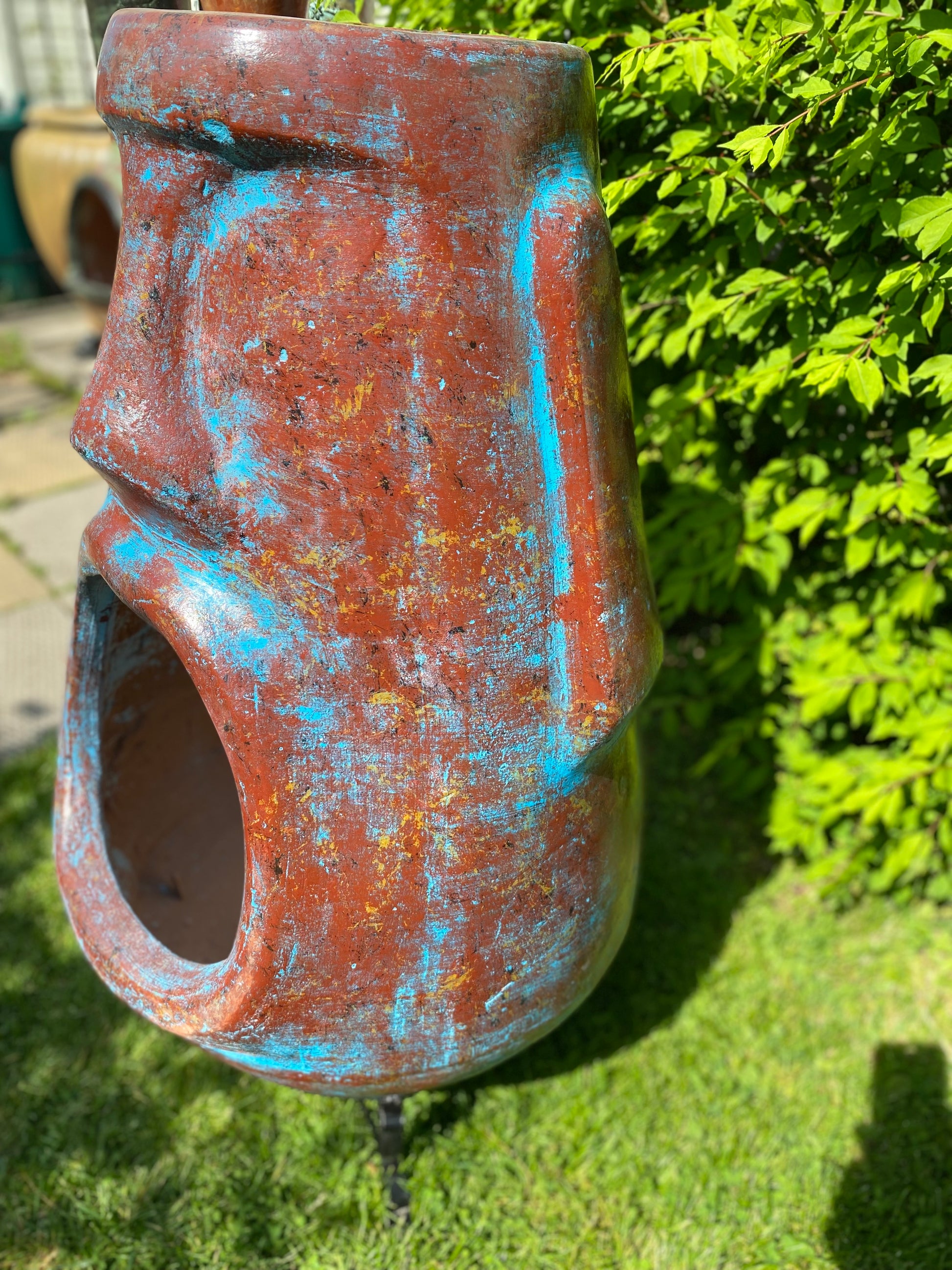 A rustic, rust-colored clay chiminea shaped like an Easter Island Moai head statue. The monolithic stone-face design features a prominent brow, a long straight nose, and a square chin. Flames flicker brightly from the wide, open mouth cavity, which serves as the fireplace opening. The outdoor heater sits on a black metal three-legged stand. close up - blue colour