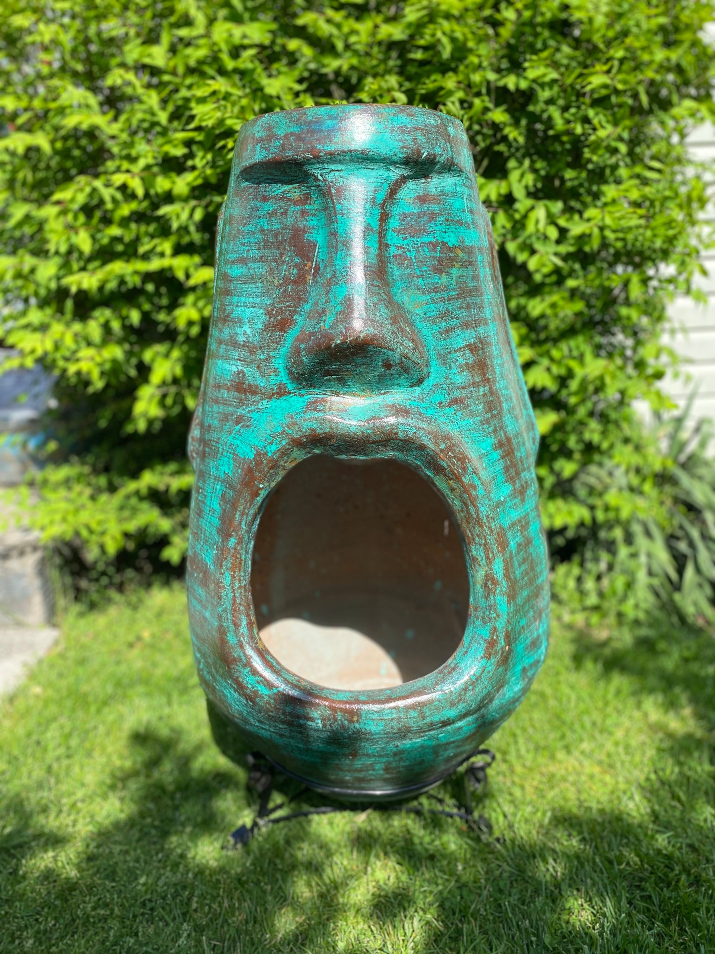 A rustic, rust-colored clay chiminea shaped like an Easter Island Moai head statue. The monolithic stone-face design features a prominent brow, a long straight nose, and a square chin. Flames flicker brightly from the wide, open mouth cavity, which serves as the fireplace opening. The outdoor heater sits on a black metal three-legged stand.