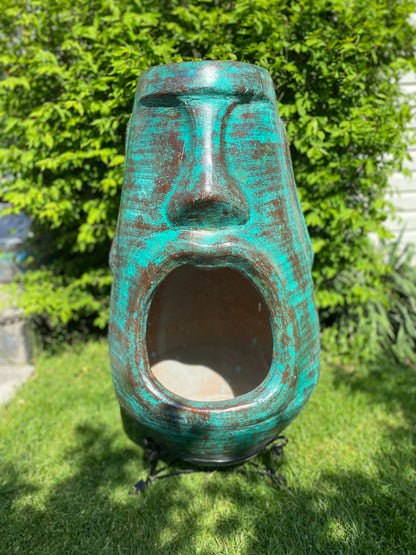 A rustic, rust-colored clay chiminea shaped like an Easter Island Moai head statue. The monolithic stone-face design features a prominent brow, a long straight nose, and a square chin. Flames flicker brightly from the wide, open mouth cavity, which serves as the fireplace opening. The outdoor heater sits on a black metal three-legged stand.
