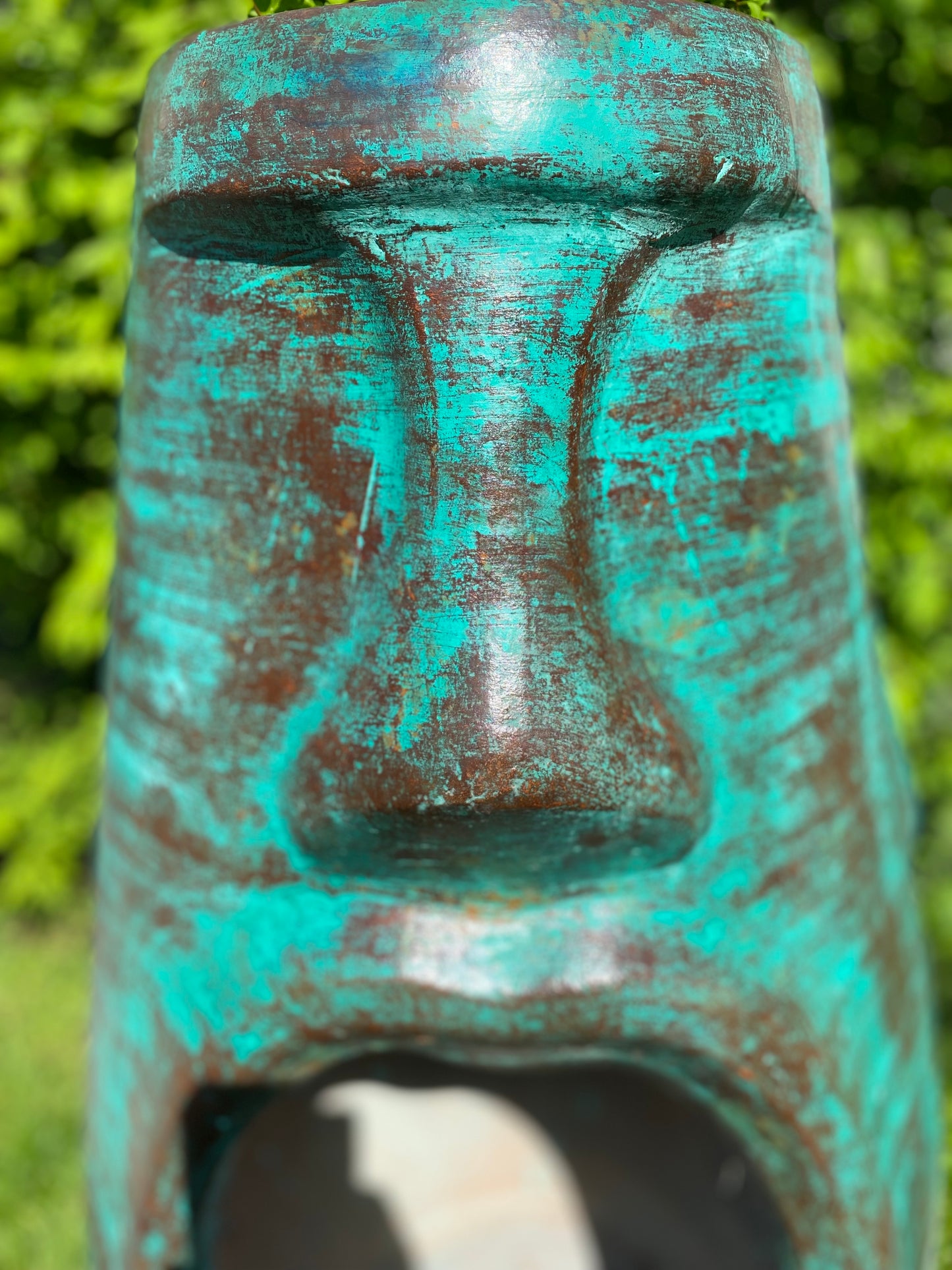 A rustic, rust-colored clay chiminea shaped like an Easter Island Moai head statue. The monolithic stone-face design features a prominent brow, a long straight nose, and a square chin. Flames flicker brightly from the wide, open mouth cavity, which serves as the fireplace opening. The outdoor heater sits on a black metal three-legged stand. close up green