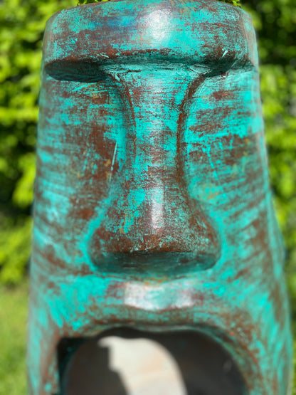 A rustic, rust-colored clay chiminea shaped like an Easter Island Moai head statue. The monolithic stone-face design features a prominent brow, a long straight nose, and a square chin. Flames flicker brightly from the wide, open mouth cavity, which serves as the fireplace opening. The outdoor heater sits on a black metal three-legged stand. close up green