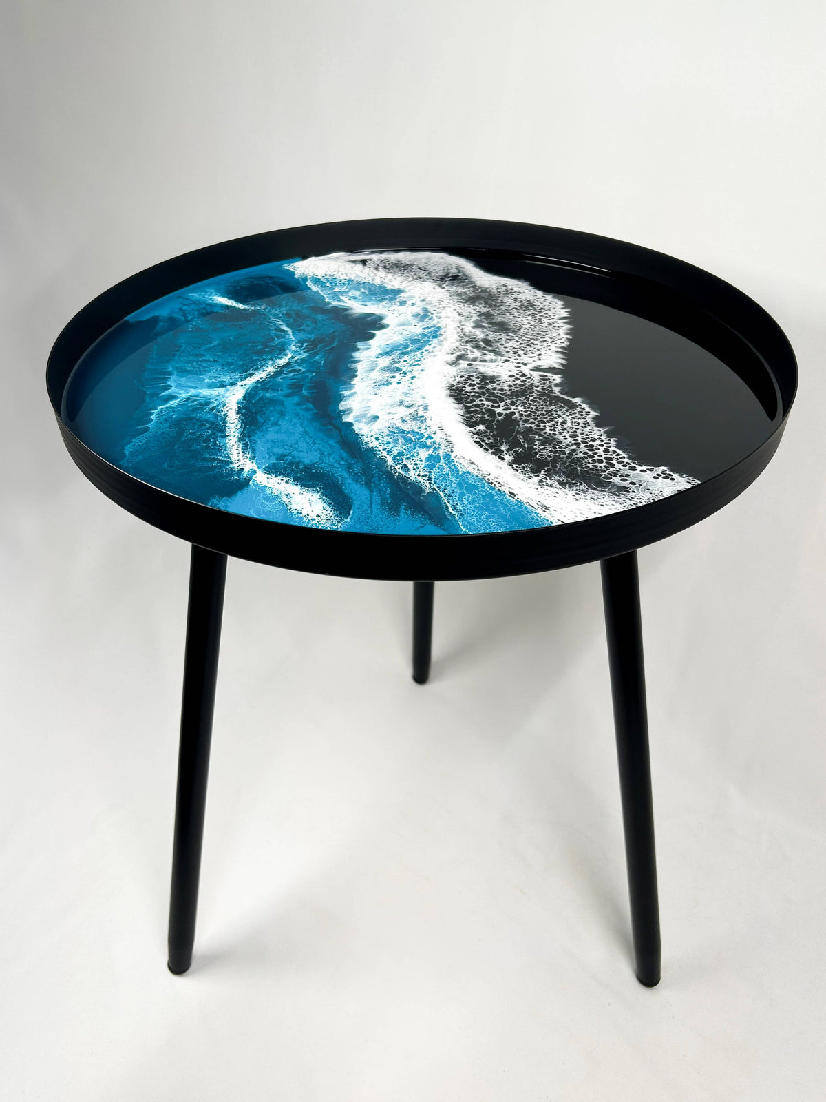 Round resin side table with blue and white ocean-wave patterns, a black rim and three black metal legs on a plain background.