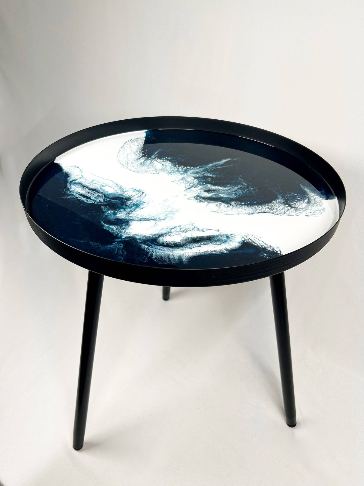 Round side table with flowing navy and white resin patterns, a black rim and three black metal legs on a plain background.
