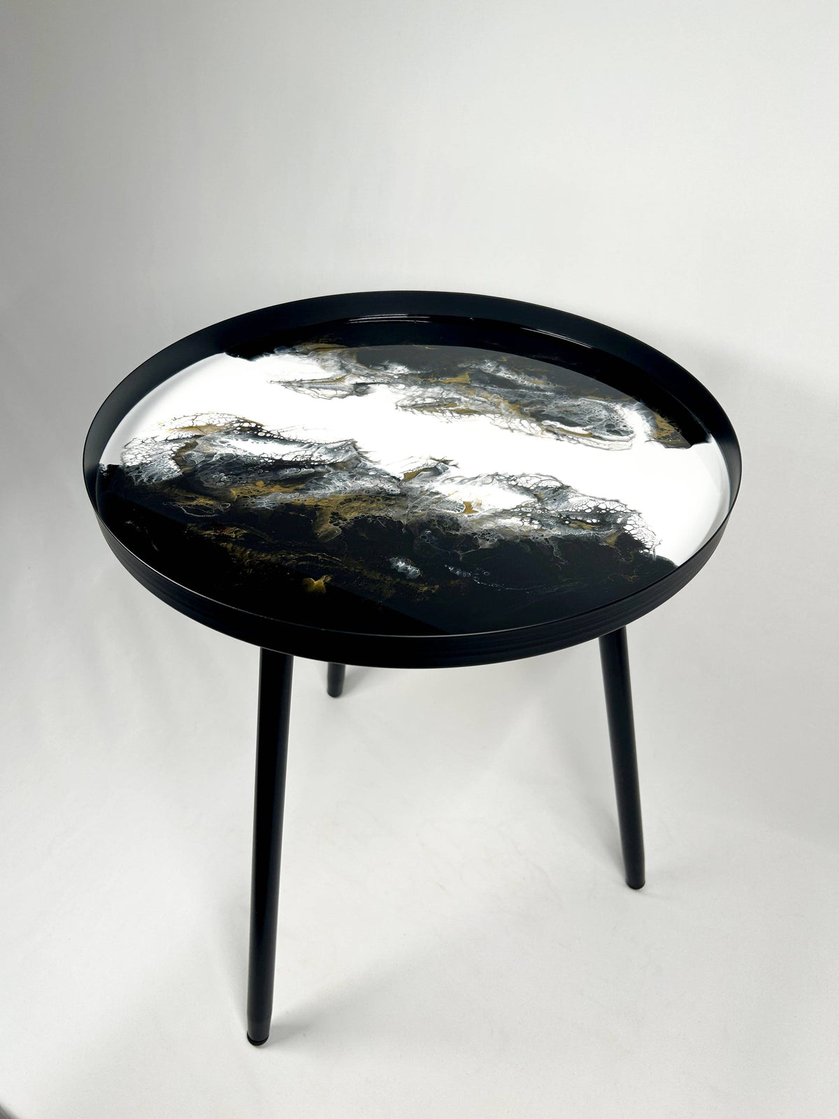 Round resin side table with black, white and gold patterns, a raised black rim and three black metal legs.